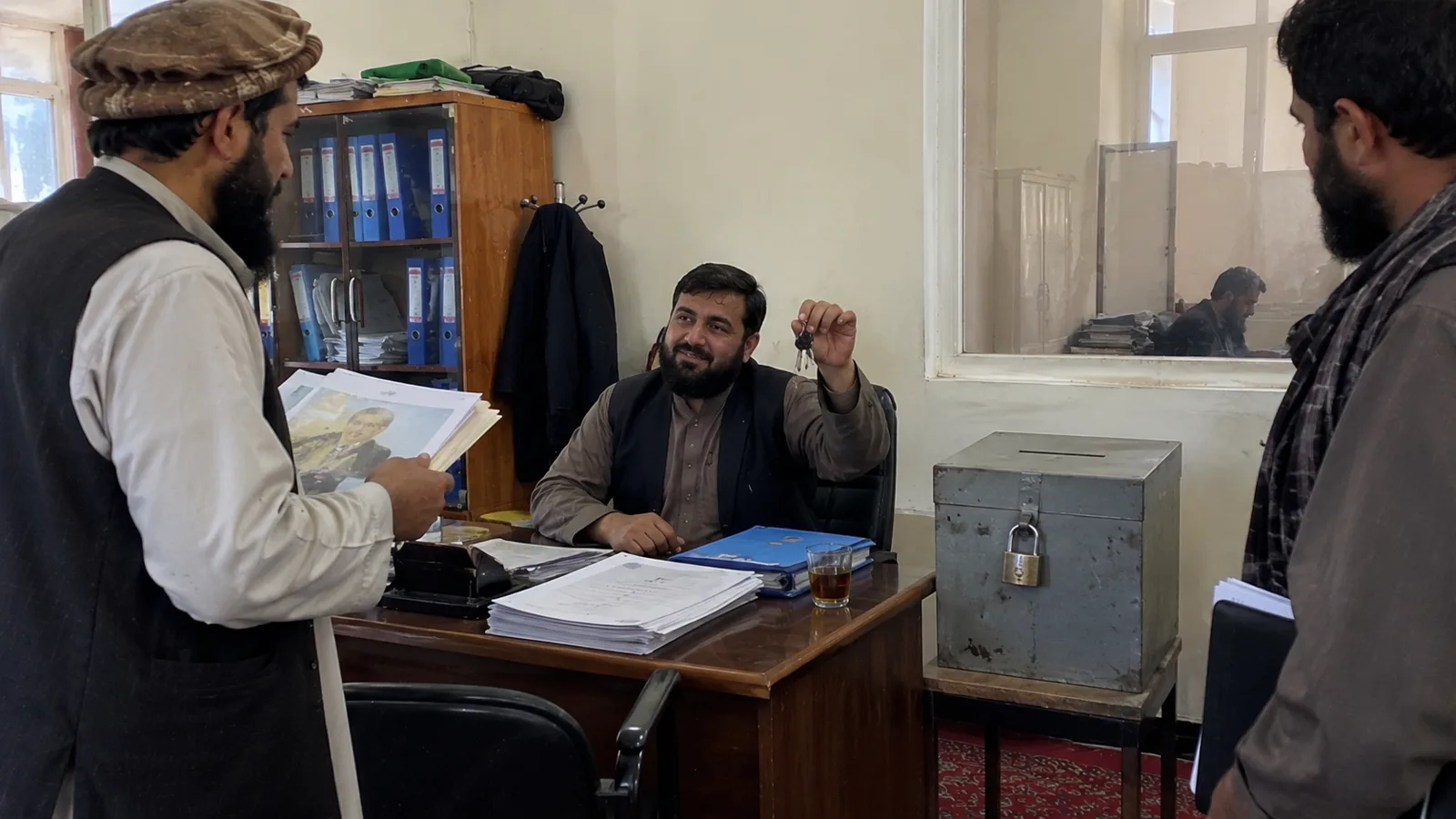 A seated man holds a key beside a locked complaints box on an office desk.
