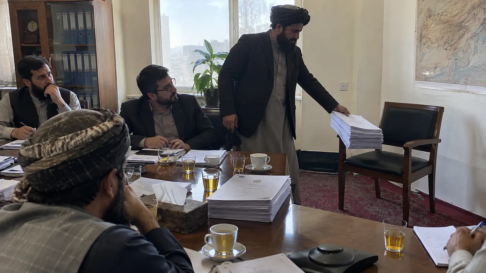 Men sit around a meeting table covered with papers and tea glasses.